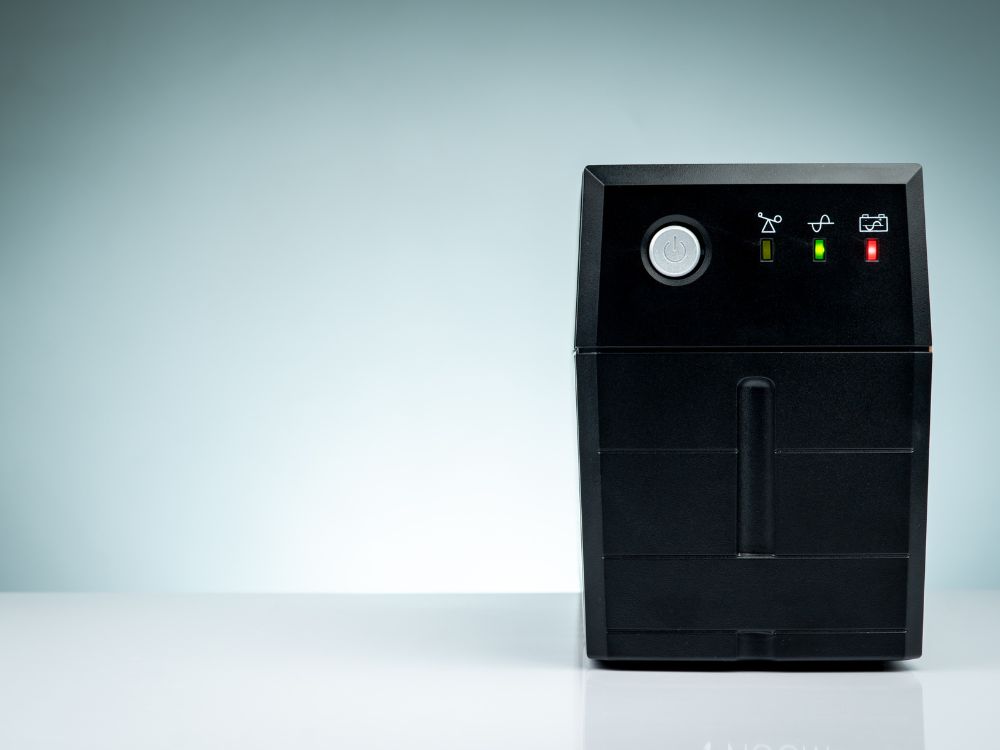 Estabilizador de energia preto com luzes indicadoras acesas, usado para proteger e regular a tensão de equipamentos eletrônicos.