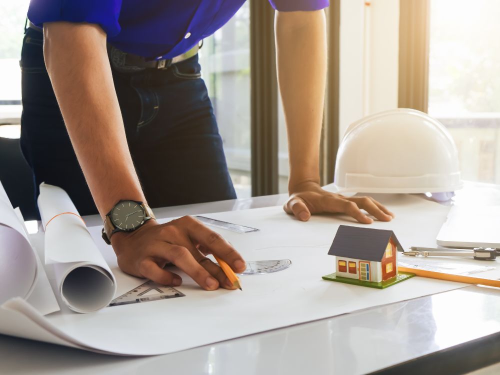 Profissional da área de arquitetura analisando projeto sobre a mesa, com plantas técnicas, régua, lápis, capacete de segurança e maquete de casa, representando o planejamento e desenvolvimento de projetos arquitetônicos.