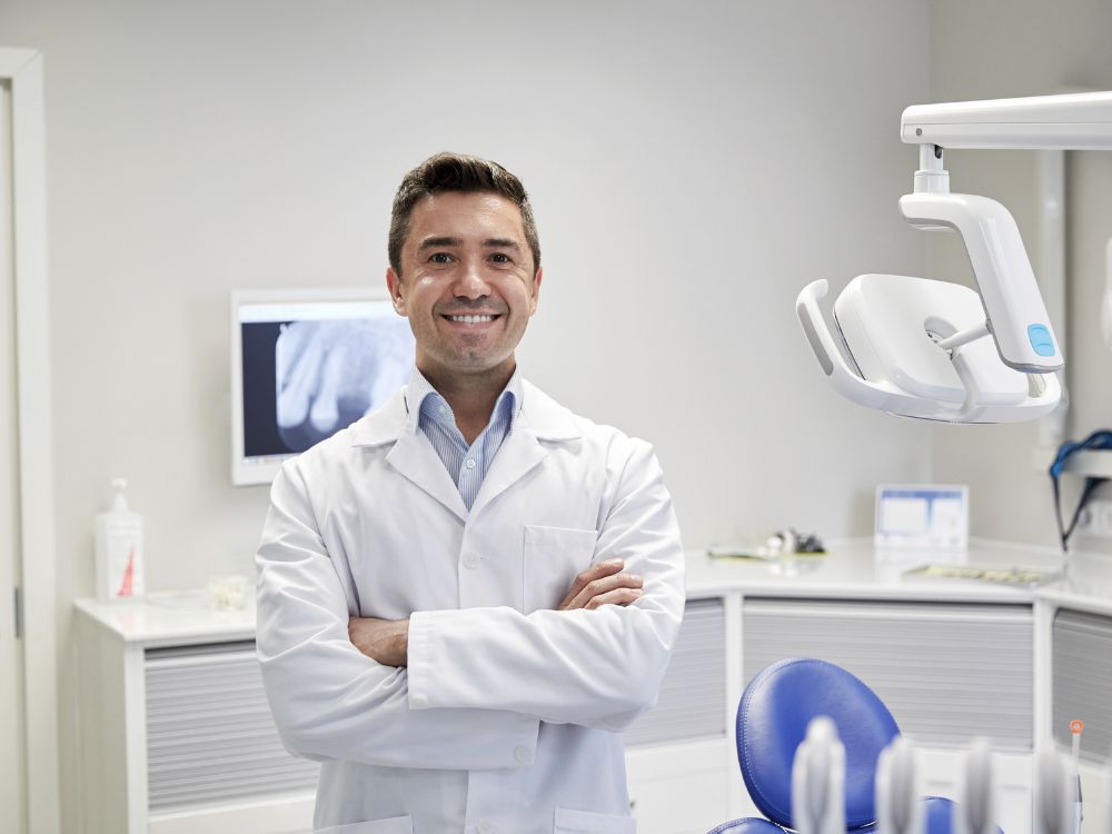 Dentista sorrindo em consultório odontológico com equipamentos ao fundo e cadeira de atendimento pronta.