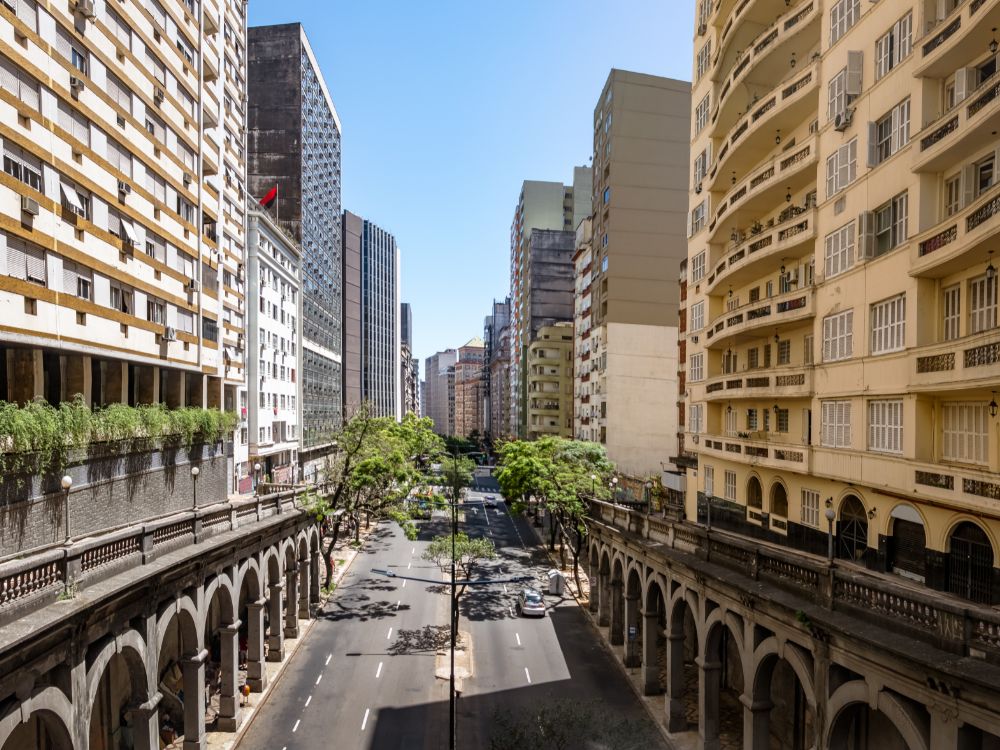 Avenida urbana cercada por prédios residenciais e comerciais, com calçadas arborizadas e arquitetura com arcos em destaque.