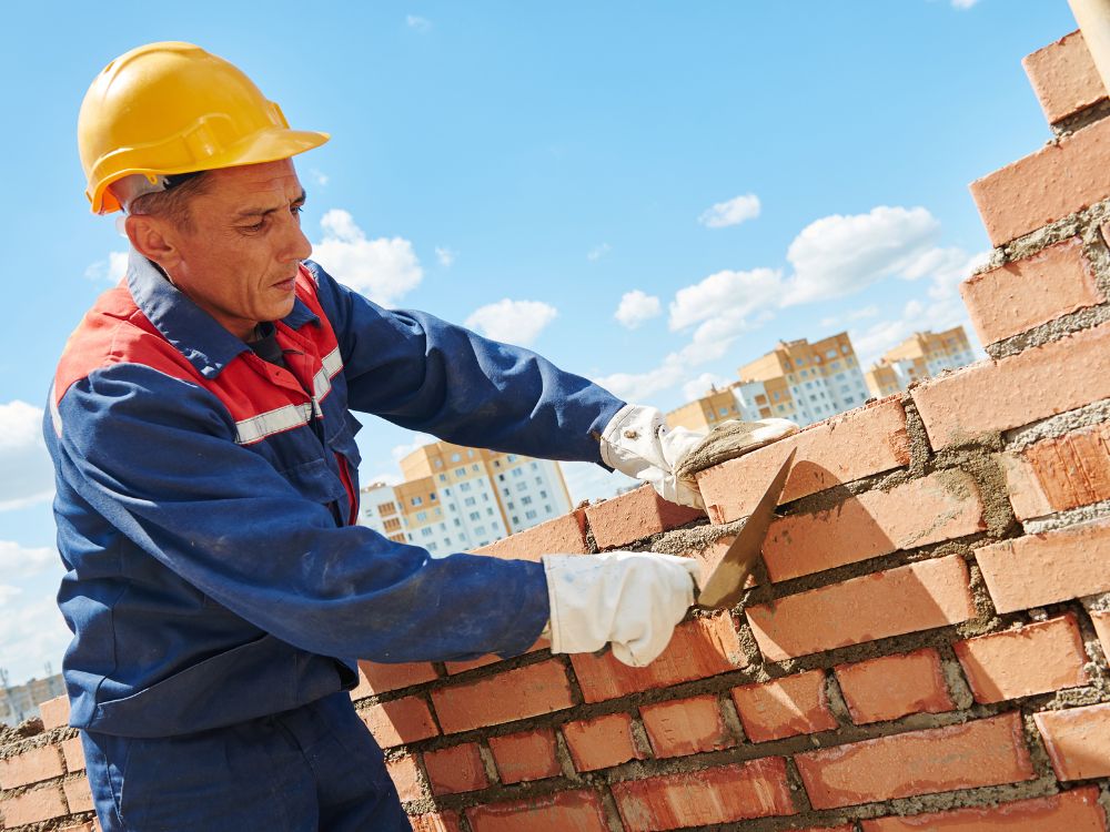 Pedreiro com capacete e luvas assentando tijolos com colher de pedreiro em construção ao ar livre.