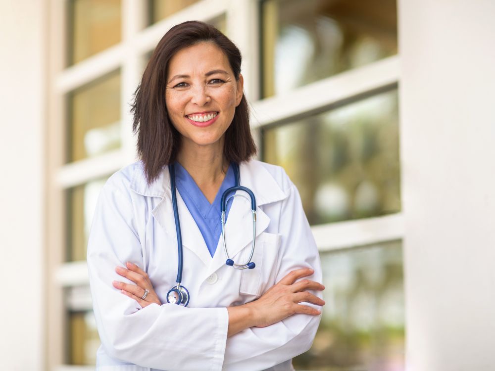 Médica com estetoscópio e jaleco branco sorrindo em ambiente clínico, representando o que faz um ginecologista endocrinologista no cuidado da saúde hormonal feminina.