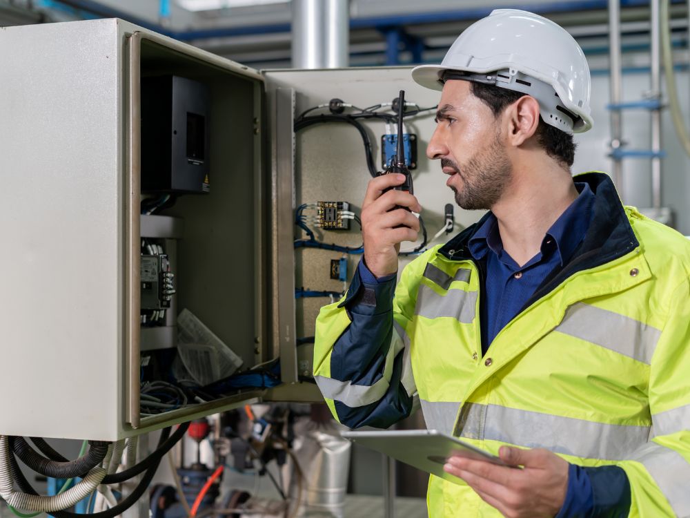 Mecânico de manutenção industrial analisando painel elétrico com rádio comunicador e tablet, realizando inspeção técnica e monitoramento de equipamentos industriais.